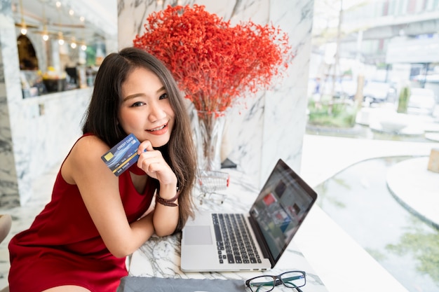 Photo jeune jolie femme asiatique assise souriant à la recherche d'appareil photo à l'aide d'une carte de crédit pour effectuer une transaction de paiement lors de vos achats sur la boutique en ligne au café