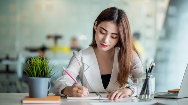 Jeune jolie femme asiatique travaillant sur ordinateur portable et prendre des notes alors qu'il était assis à la table au bureau