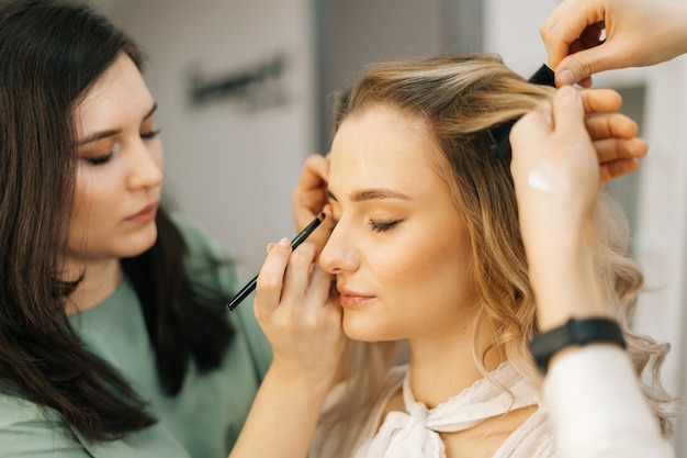 Jeune jolie femme assise sur une chaise et bénéficiant d'un service de maquillage professionnel d'une maquilleuse