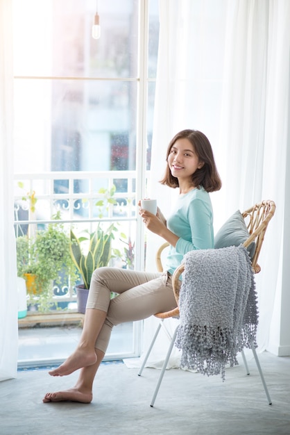 Jeune jolie femme assise à la fenêtre ouverte buvant du café et regardant à l'extérieur profite du repos