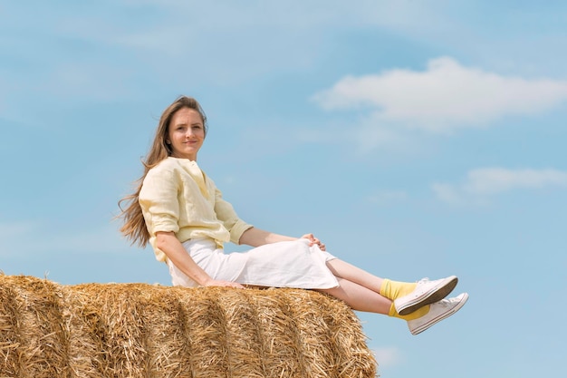 Jeune jolie femme assise sur des meules de foin sur fond de ciel bleu