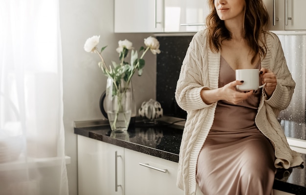 Jeune jolie femme assise sur une table tenant une tasse de café chaud dans la salle de cuisine avec un intérieur moderne blanc