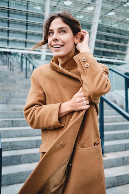 Jeune jolie femme attachant le manteau du froid joyeusement regardant loin marchant en plein air