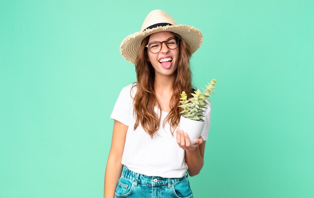 Jeune jolie femme à l'attitude joyeuse et rebelle, plaisantant et tirant la langue avec un chapeau de paille et tenant un cactus