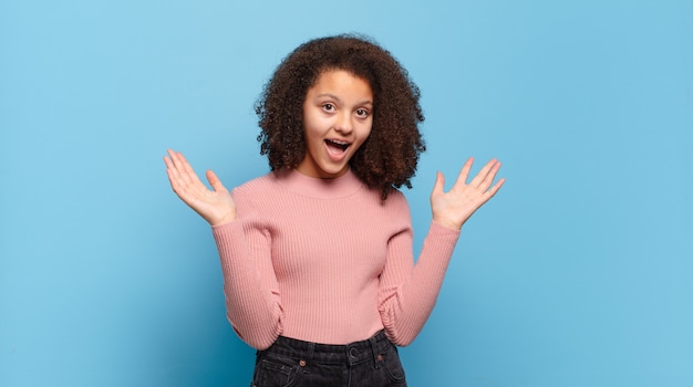 Jeune jolie femme aux cheveux afro et pull rose posant sur le mur bleu