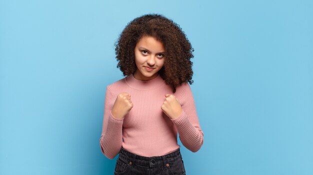 Jeune jolie femme aux cheveux afro et pull rose posant sur le mur bleu