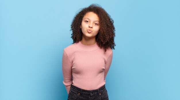 Jeune jolie femme aux cheveux afro et pull rose posant sur le mur bleu