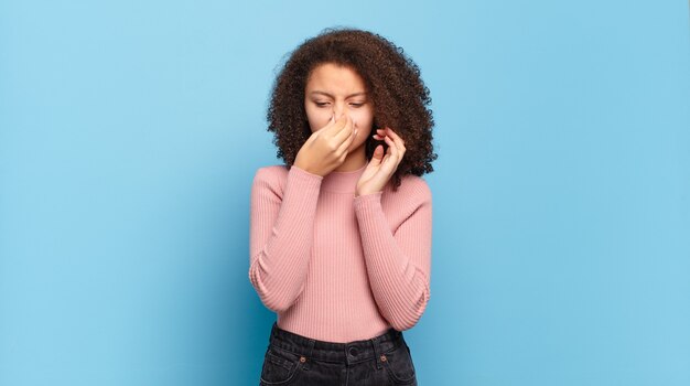 Jeune jolie femme aux cheveux afro et pull rose posant sur le mur bleu