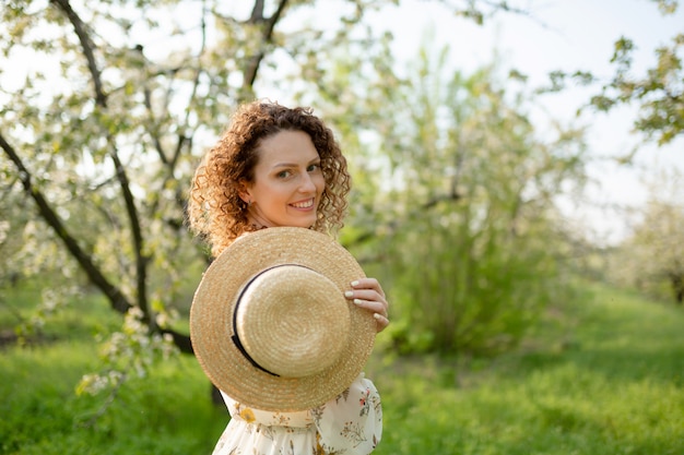 Jeune jolie femme aux cheveux bouclés dans un élégant chapeau en osier se promène dans un jardin vert en fleurs au printemps