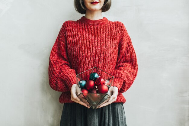 Jeune jolie femme aux lèvres rouges, pull en tricot rouge et jupe grise tenant dans ses mains vase géométrique avec des jouets de Noël rouge et bleu