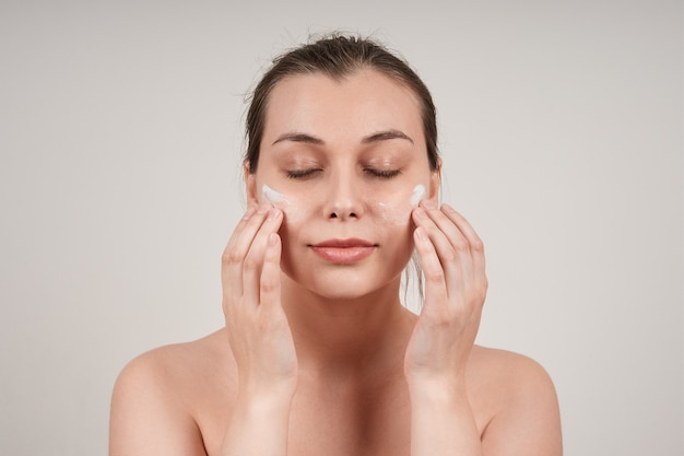 Jeune jolie femme aux seins nus appliquant une crème nourrissante au visage, les yeux fermés, isoler sur un mur gris.