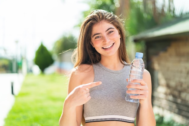 Une jeune et jolie femme blanche avec une bouteille d'eau à l'extérieur et le pointant