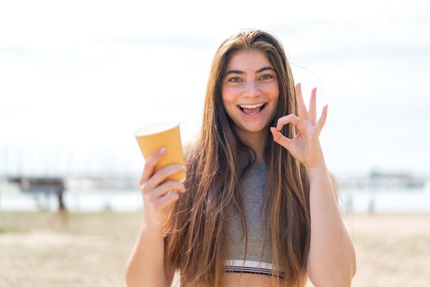 Une jeune et jolie femme blanche tenant un café à emporter à l'extérieur montrant un signe ok avec les doigts