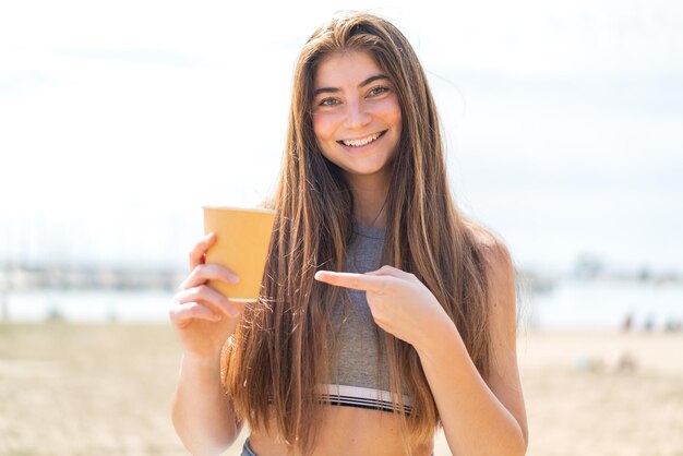 Une jeune et jolie femme blanche tenant un café à emporter à l'extérieur et le pointant