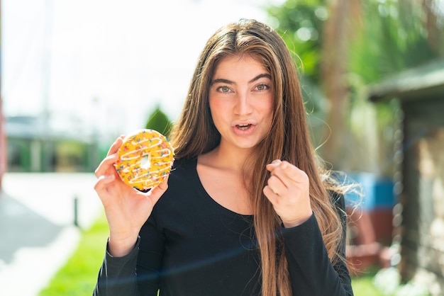 Une jeune et jolie femme blanche tenant un donut à l'extérieur étonnée et pointant vers l'avant