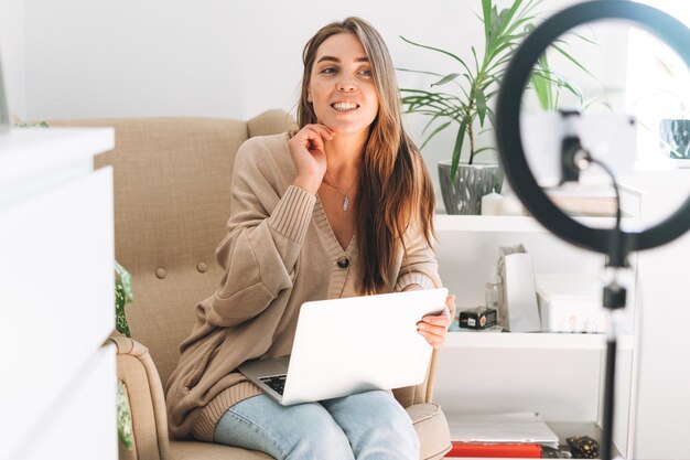 Jeune jolie femme blogueuse enregistre une vidéo sur un téléphone portable à l'aide d'une lampe annulaire assise sur une chaise avec un ordinateur portable dans la chambre avec des plantes vertes à la maison