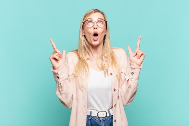 Jeune jolie femme blonde à l'air choquée étonnée et la bouche ouverte pointant vers le haut avec les deux mains pour copier l'espace