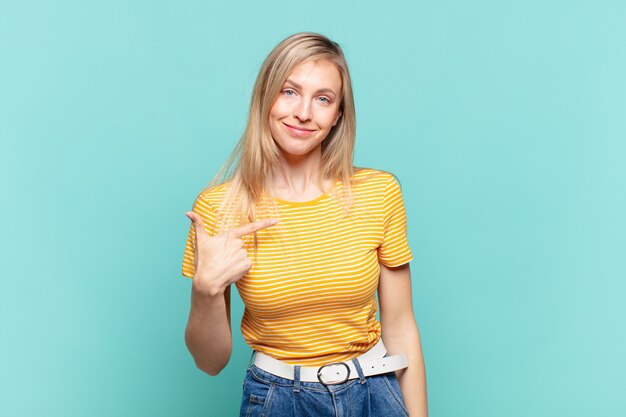 Jeune jolie femme blonde à l'air fière, confiante et heureuse, souriante et se montrant elle-même ou faisant le signe numéro un