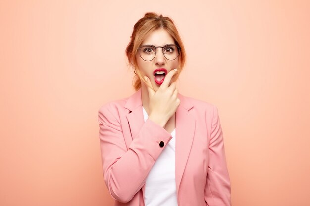 Jeune jolie femme blonde avec la bouche et les yeux grands ouverts et la main sur le menton, se sentant désagréablement choquée