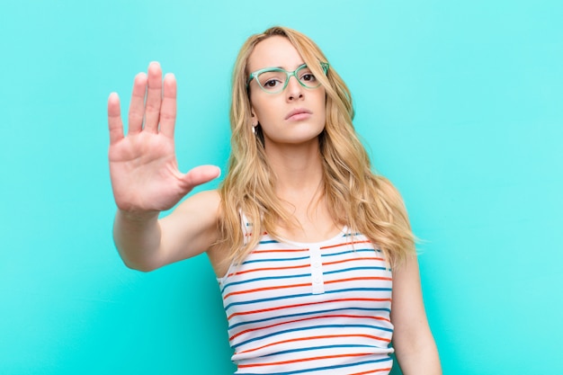 Jeune jolie femme blonde à la grave, sévère, mécontente et en colère montrant la paume ouverte faisant le geste d'arrêt contre le mur de couleur plat