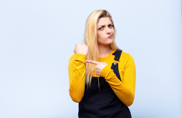 Photo jeune jolie femme blonde à l'impatience et en colère, montrant la montre, demandant la ponctualité, veut être à l'heure contre le mur plat