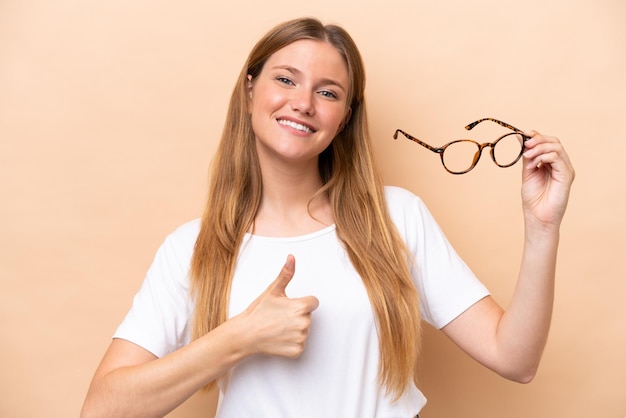 Jeune jolie femme blonde avec des lunettes isolées sur fond beige avec les pouces vers le haut parce que quelque chose de bien s'est produit