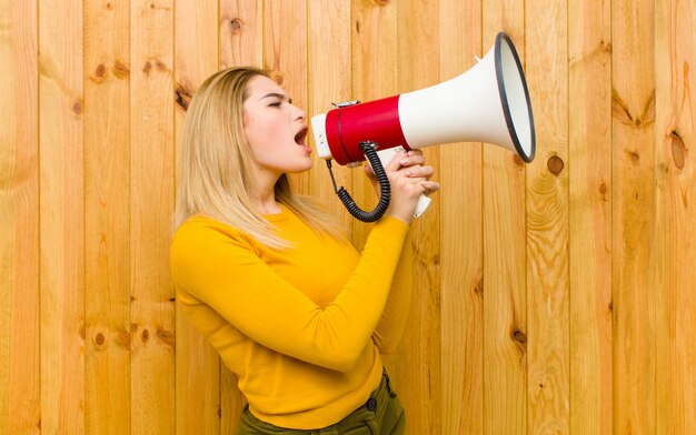 Jeune jolie femme blonde avec un mégaphone contre un mur en bois