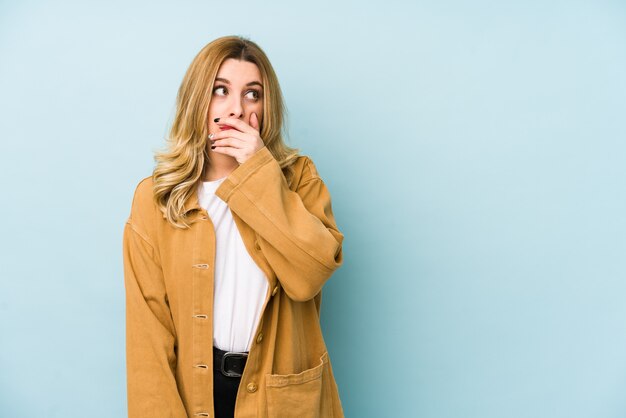 Jeune jolie femme blonde réfléchie à la recherche d'un espace vide couvrant la bouche avec la main.