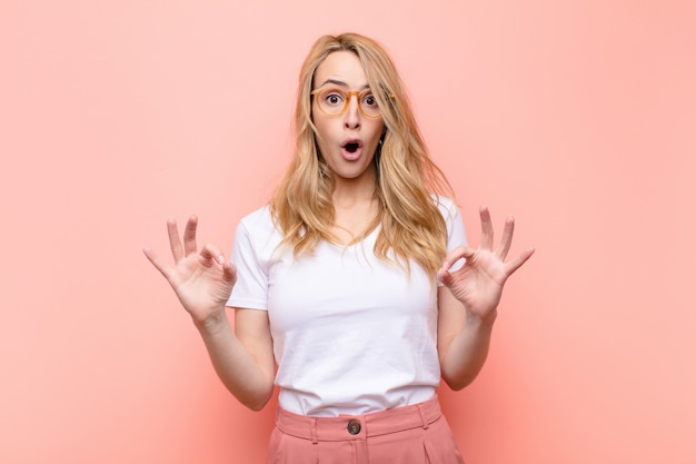 Jeune jolie femme blonde se sentant choquée, étonnée et surprise, montrant l'approbation faisant signe correct avec les deux mains contre le mur de couleur plat