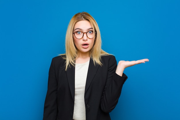 Jeune jolie femme blonde semblant surprise et choquée, avec la mâchoire tombée, tenant un objet avec une main ouverte sur le côté contre le mur plat