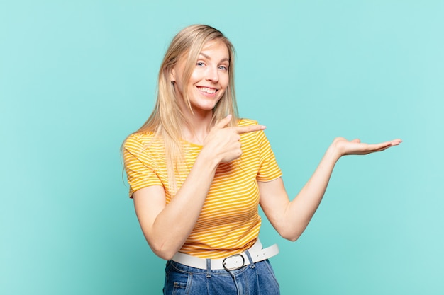 Jeune jolie femme blonde souriant joyeusement et pointant pour copier l'espace sur la paume sur le côté, montrant ou faisant la publicité d'un objet