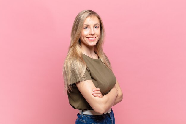 Jeune jolie femme blonde souriante à la caméra avec les bras croisés et une expression heureuse, confiante et satisfaite, vue latérale