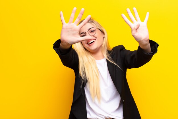 Jeune jolie femme blonde souriante et à la sympathique, montrant le numéro neuf ou neuvième avec la main en avant, compte à rebours contre le mur jaune