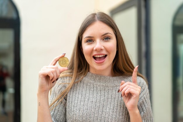 Jeune jolie femme blonde tenant un Bitcoin à l'extérieur pointant vers le haut une bonne idée