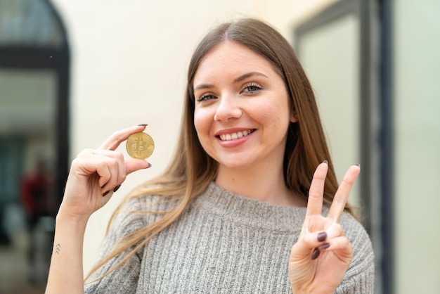 Jeune jolie femme blonde tenant un Bitcoin à l'extérieur souriant et montrant le signe de la victoire