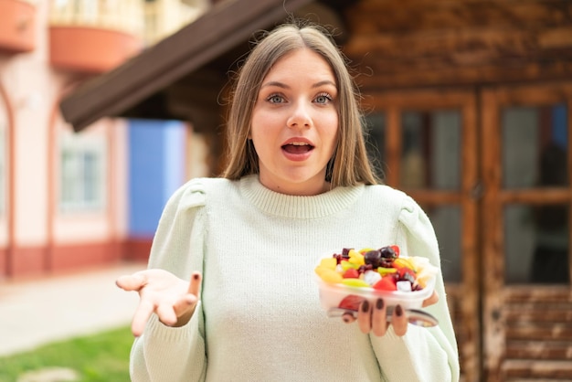 Jeune jolie femme blonde tenant un bol de fruits à l'extérieur avec une expression faciale choquée