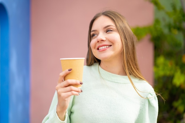 Jeune jolie femme blonde tenant un café à emporter à l'extérieur en levant les yeux en souriant
