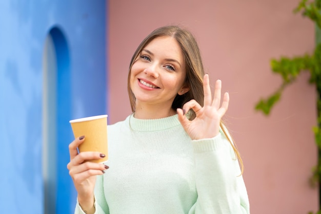 Jeune jolie femme blonde tenant un café à emporter à l'extérieur montrant un signe ok avec les doigts