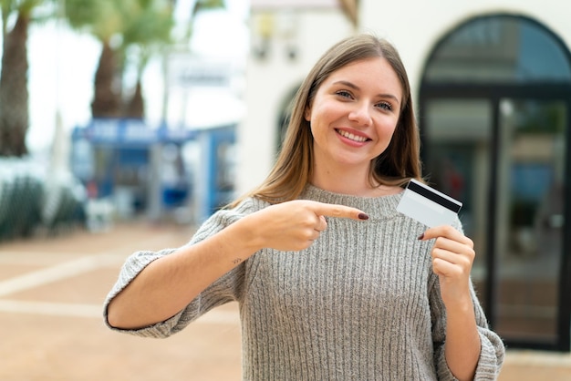 Jeune jolie femme blonde tenant une carte de crédit à l'extérieur et la pointant