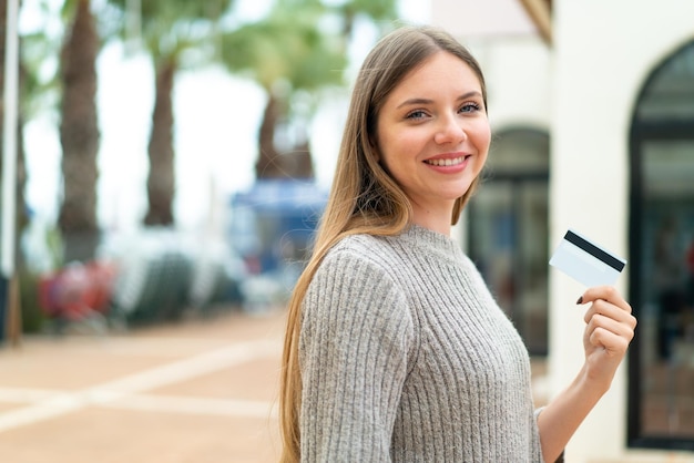 Jeune jolie femme blonde tenant une carte de crédit à l'extérieur souriant beaucoup