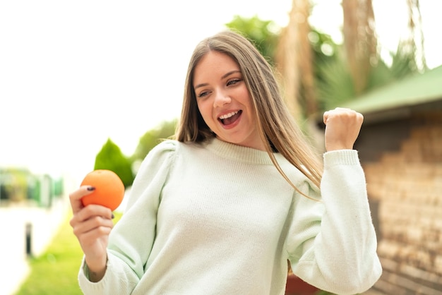 Jeune jolie femme blonde tenant une orange à l'extérieur célébrant une victoire