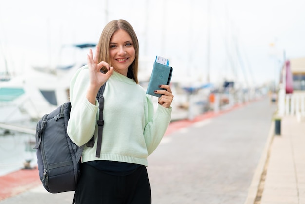 Jeune jolie femme blonde tenant un passeport à l'extérieur montrant un signe ok avec les doigts