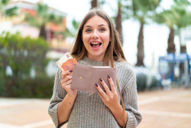 Jeune jolie femme blonde tenant un portefeuille avec de l'argent avec une expression heureuse