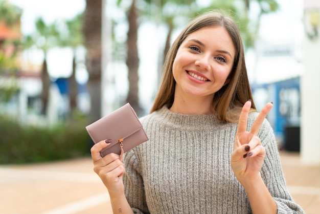 Jeune jolie femme blonde tenant un portefeuille à l'extérieur souriant et montrant le signe de la victoire