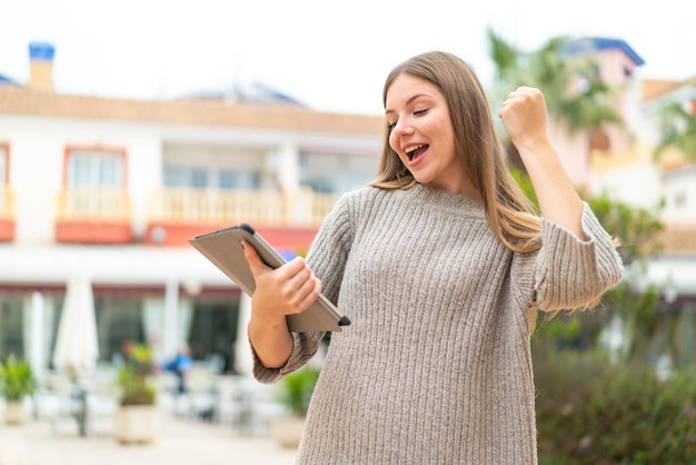 Jeune jolie femme blonde tenant une tablette à l'extérieur célébrant une victoire