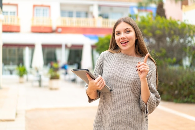 Jeune jolie femme blonde tenant une tablette à l'extérieur pointant vers le haut une bonne idée