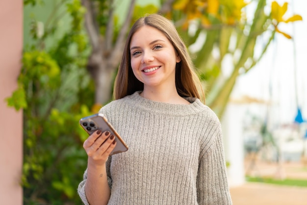 Jeune jolie femme blonde utilisant un téléphone portable à l'extérieur souriant beaucoup