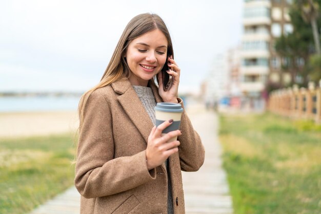 Jeune jolie femme blonde utilisant un téléphone portable et tenant un café avec une expression heureuse