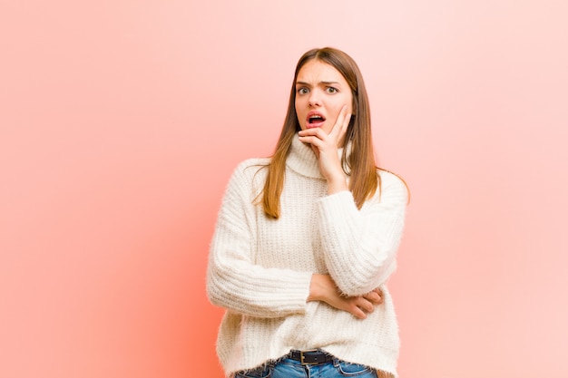 Jeune jolie femme bouche bée sous le choc et l'incrédulité, la main sur la joue et le bras croisés, se sentant stupéfaite et stupéfaite contre le mur rose