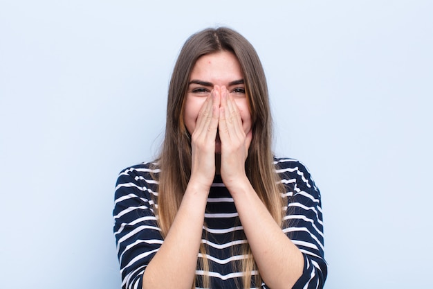 Jeune jolie femme à la bouche heureuse, gaie, chanceuse et surprise couvrant les deux mains contre le mur bleu doux